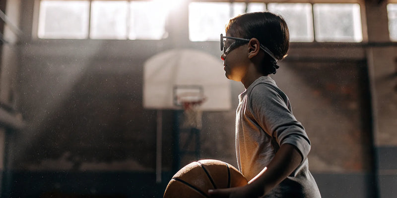Kid wearing protective basketball glasses holding a basketball on a court.
